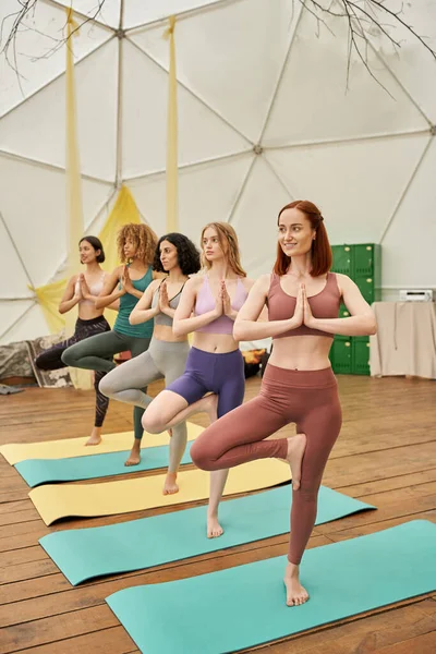 Positive woman practicing yoga in tree pose and looking away near multiethnic girlfriends — Stock Photo