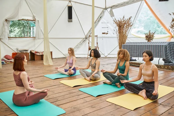 Novias multiétnicas meditando en pose de loto en colchonetas de yoga en centro de retiro moderno - foto de stock