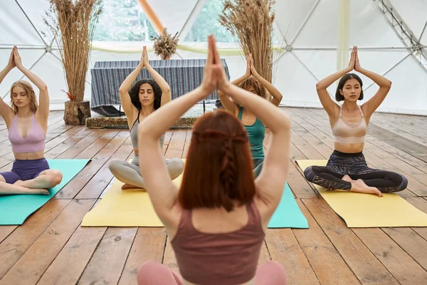 Yoga coach showing asana with raised praying hands to multiethnic women meditating in retreat center — Stock Photo