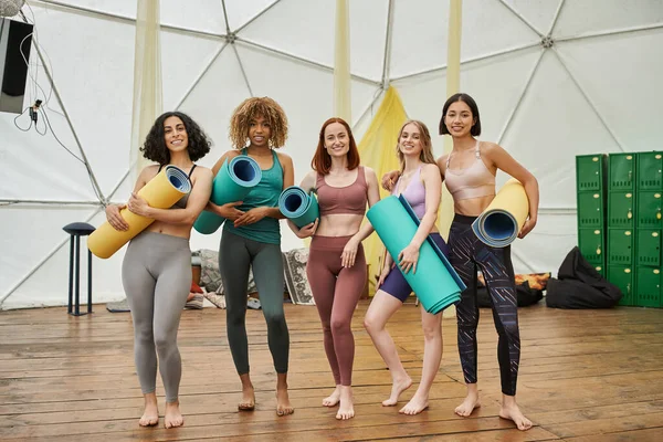Comprimento total de mulheres multiétnicas felizes em sportswear de pé com tapetes de ioga e olhando para a câmera — Fotografia de Stock