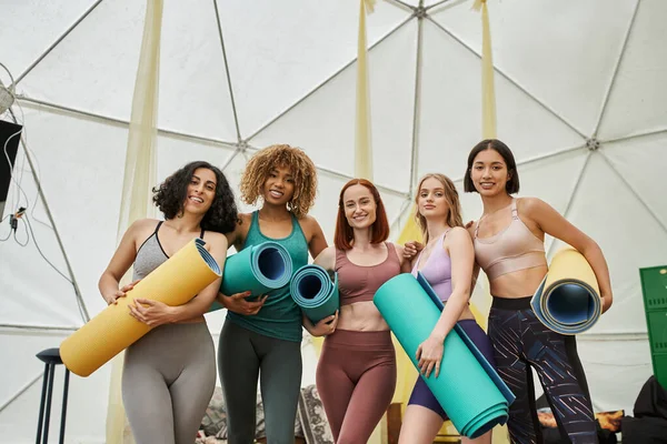 Novias multiétnicas con colchonetas de yoga sonriendo a la cámara en el centro de retiro moderno - foto de stock