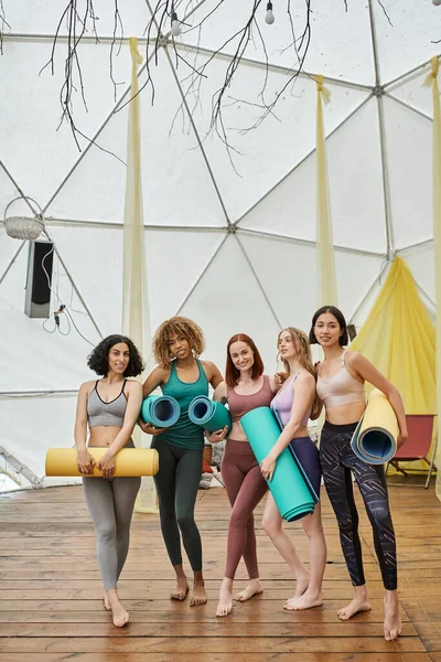 Comprimento total de mulheres multiétnicas em sportswear de pé com tapetes de ioga no centro de retiro moderno — Fotografia de Stock