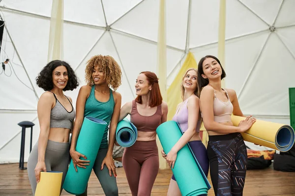 Novias multiétnicas en ropa deportiva sosteniendo esteras de yoga y riendo en el centro de retiro moderno - foto de stock