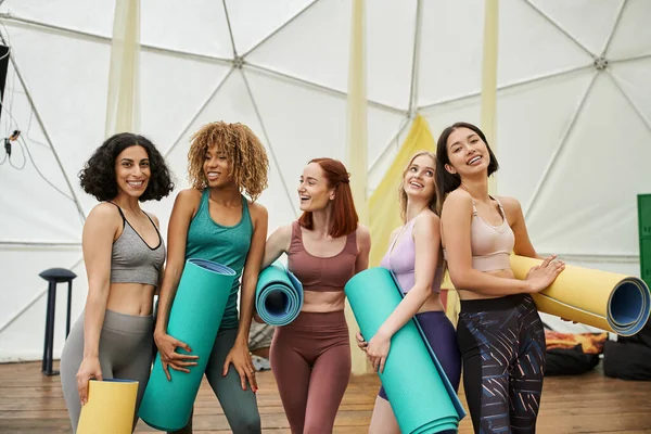 Multiethnic girlfriends in sportswear holding yoga mats and laughing in modern retreat center — Stock Photo