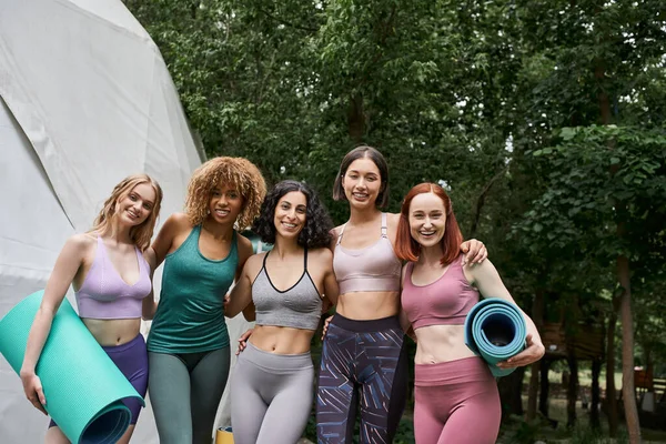Amigas multiétnicas positivas en ropa deportiva sosteniendo esteras de yoga y sonriendo a la cámara, parque - foto de stock