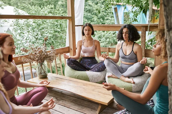 Multiethnic girlfriends in sportswear meditating in lotus pose in patio of retreat center — Stock Photo