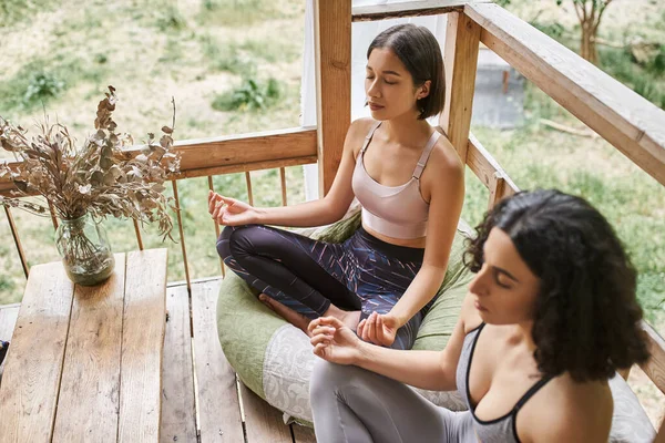Young woman meditating in lotus pose near multiracial girlfriend in patio of retreat center — Stock Photo