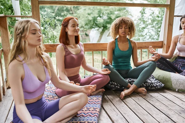 Multiethnic girlfriends with closed eyes meditating in lotus pose in cottage on retreat center — Stock Photo