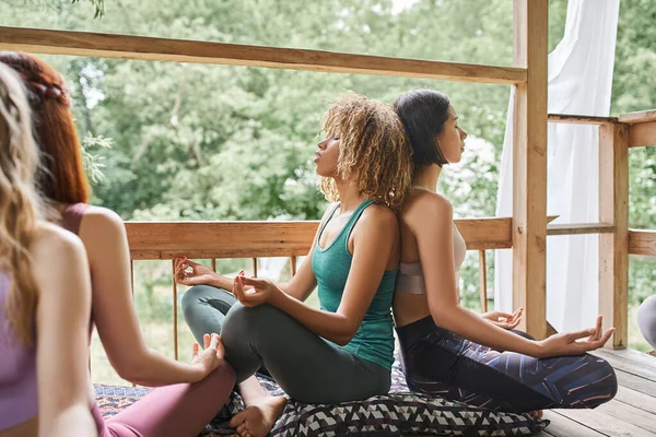 Novias multiétnicas meditando en la pose de loto y sentado espalda con espalda en el patio de la casa acogedora - foto de stock