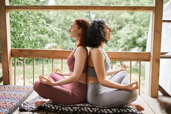 Carefree girlfriends in sportswear meditating in lotus pose with closed eyes, retreat center, patio — Stock Photo