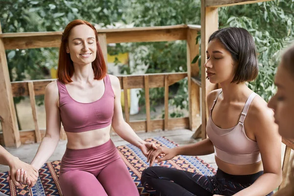 Cheerful woman holding hands with girlfriends and meditating with closed eyes in retreat center — Stock Photo
