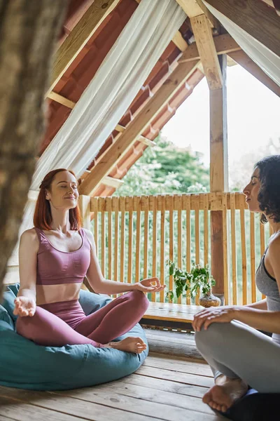 Smiling woman with closed eyes sitting in lotus pose near multiracial girlfriend in cozy patio — Stock Photo