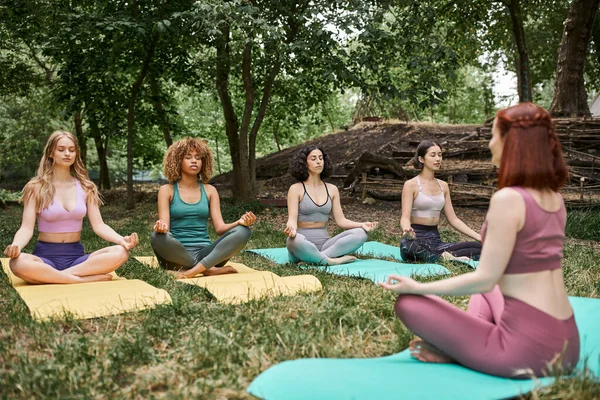 Mulheres multiétnicas em tapetes de ioga meditando em pose de lótus perto de treinador no parque — Fotografia de Stock
