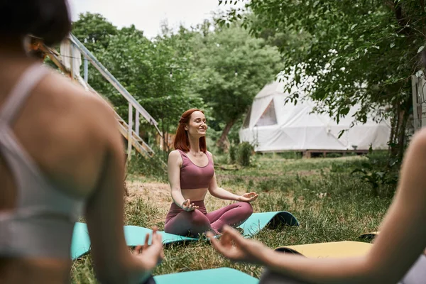 Carefree woman meditating in lotus pose near blurred girlfriends in outdoor retreat center — Stock Photo