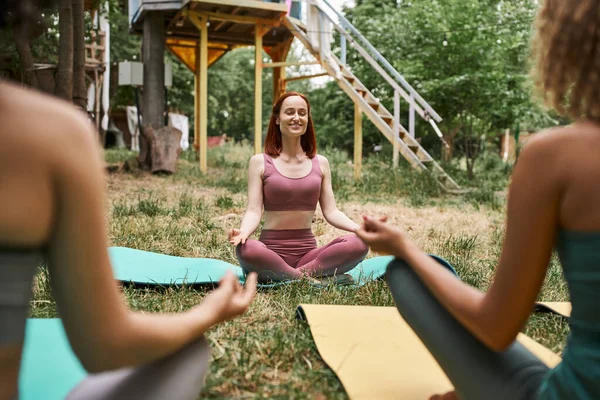 Веселая женщина с закрытыми глазами медитирует в парке ретритного центра рядом с размытыми подругами — стоковое фото