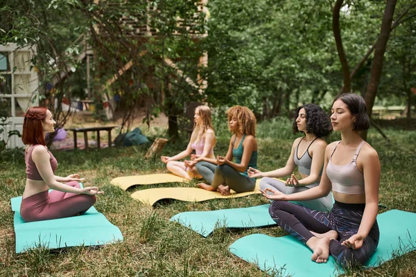 Многонациональные девушки в спортивной одежде медитируют в позе лотоса в парке, внутренний мир, гармония — стоковое фото