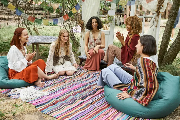 Laughing girlfriends in boho style outfit talking on colorful blanket in park of retreat center — Stock Photo