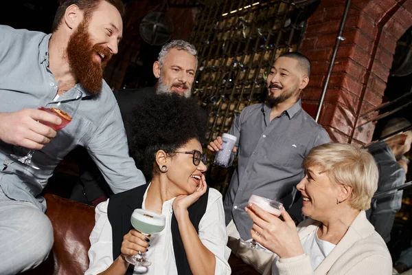 Femmes multiethniques heureuses avec des cocktails parlant près des hommes barbus dans le bar, amusement et loisirs après le travail — Photo de stock