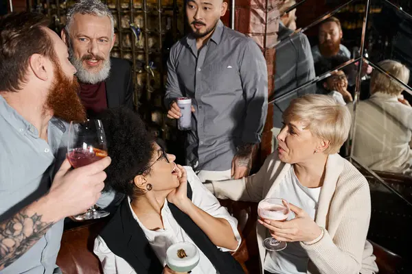 Hommes barbus parlant près de femmes émerveillées, collègues multiethniques avec des cocktails relaxant dans le bar — Photo de stock