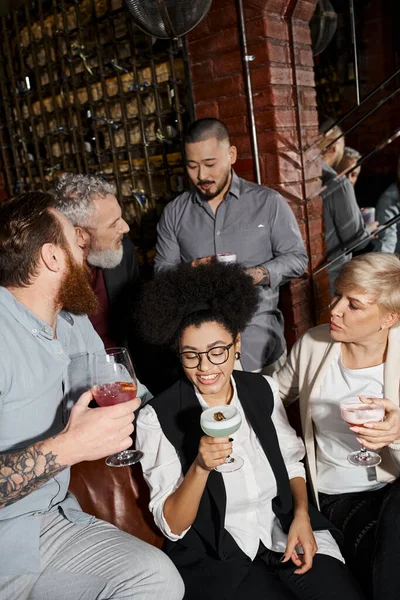 Bearded tattooed men talking near women drinking cocktails in bar, leisure of multiethnic colleagues — Stock Photo