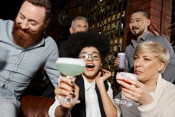 Вне себя от радости африканская американка с коктейлем, смотрящая в камеру возле мультиэтнических коллег в баре — стоковое фото