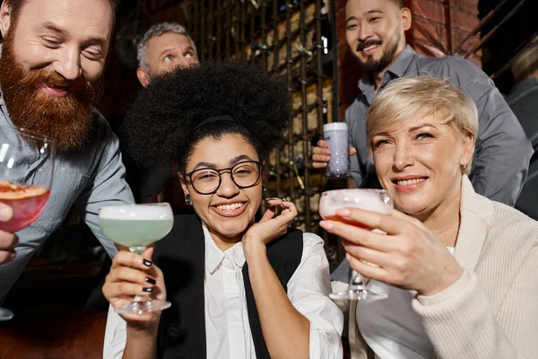 Happy women with cocktails near bearded men in bar, multiethnic colleagues resting in bar after work — Stock Photo