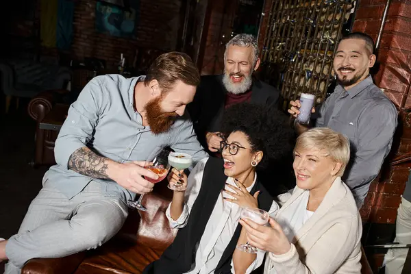 Barbudo hombre tatuado tintineo gafas con mujeres alegres cerca de amigos multiétnicos en bar de cócteles - foto de stock