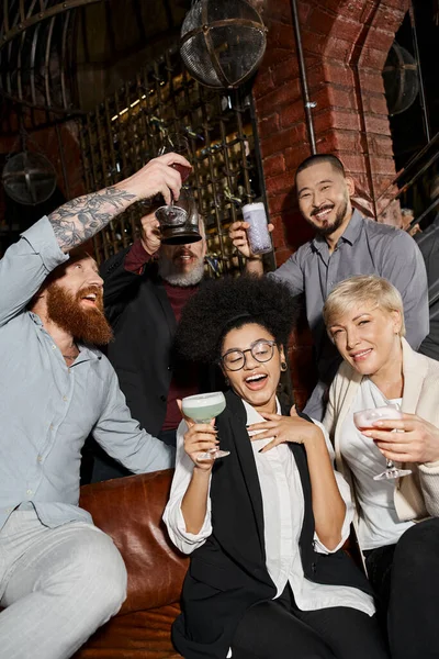 Barbudos hombres tatuados tostadas cerca de mujeres despreocupadas en el bar de cócteles, relajarse de colegas multiétnicos - foto de stock