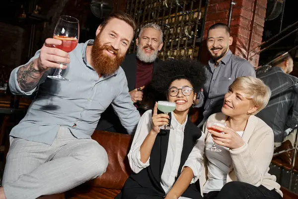 Бородатий татуйований чоловік з коктейлем дивиться на камеру поруч з багатоетнічними друзями, розважаючись у барі — стокове фото