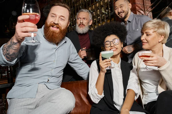 Pleased tattooed and bearded man looking at cocktail near multiethnic colleagues with glasses in bar — Stock Photo