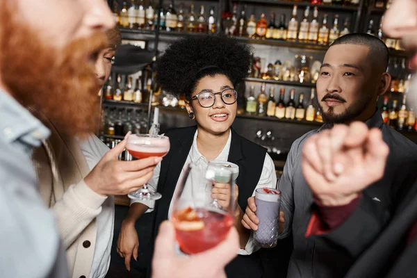 Positive african american woman holding cocktail and smiling near multiethnic work friends in bar — Stock Photo