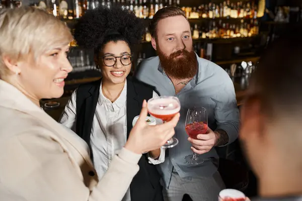 Бородатый татуированный мужчина и африканская американка улыбаются рядом с коллегами в коктейльных бокалах в баре — стоковое фото