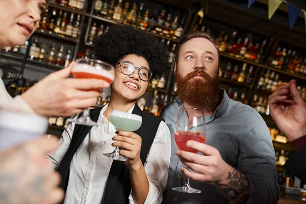 Cheerful african american woman and bearded man with cocktails resting with colleagues in bar — Stock Photo