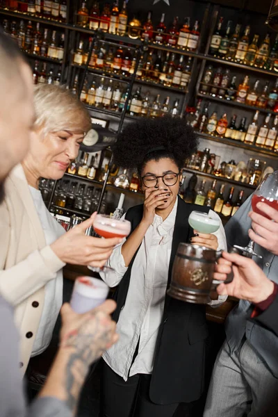 Excited african american woman laughing with closed eyes near team of colleagues with drinks in bar — Stock Photo