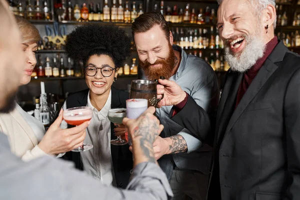 Bearded tattooed men clinking glasses with happy multiethnic women in cocktail bar after work — Stock Photo