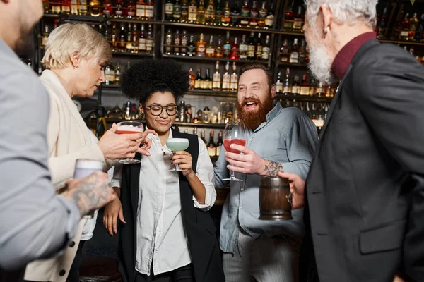 Barbudo hombre tatuado riendo cerca de amigos multiculturales sosteniendo vasos con cócteles en el bar - foto de stock