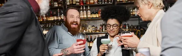 Hombre barbudo emocionado riendo cerca de amigos multiculturales sosteniendo vasos con bebidas en el bar, pancarta - foto de stock