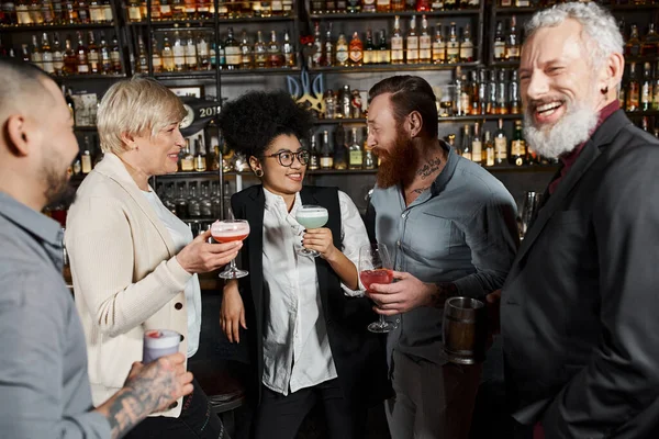 Бородатый татуированный мужчина разговаривает с улыбчивой африканской американкой рядом с коллегами по работе с напитками в баре — стоковое фото