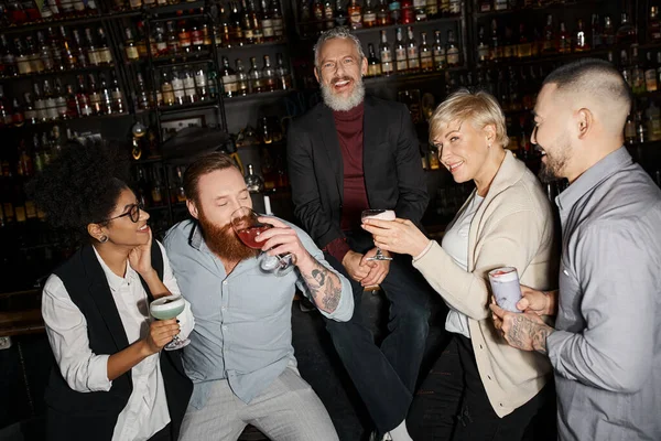Веселий бородатий чоловік дивиться на камеру біля багатоетнічних колег, п'є коктейлі в барі — стокове фото