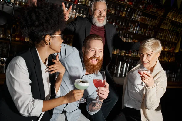Збуджений бородатий чоловік показує знак перемоги біля сміху багатоетнічних друзів, що тримають коктейлі в барі — стокове фото