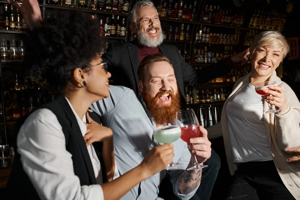 Hombres barbudos felices riendo cerca de mujeres multiculturales en el bar de cócteles, diversa fiesta de equipo después del trabajo - foto de stock