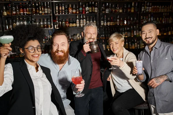 Carefree multiethnic workmates holding glasses with drinks and looking at camera in cocktail bar — Stock Photo