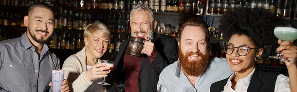 Joyful multiethnic colleagues with drinks smiling and looking at camera in cocktail bar, banner — Stock Photo