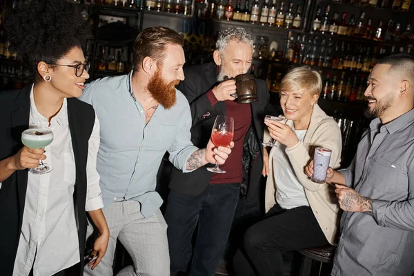 Bearded tattooed man with cocktail glass near happy multiethnic workmates in bar, fun after work — Stock Photo