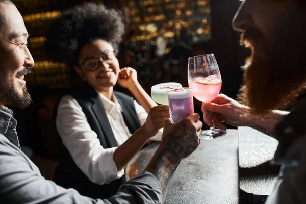 Усміхнена афроамериканка смердить коктейль окуляри з бородатими татуйованими колегами в барі — стокове фото
