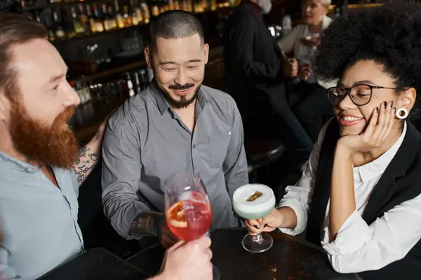 Довольная африканская американка с бородатыми татуированными коллегами, сидящая в баре с коктейлями — стоковое фото