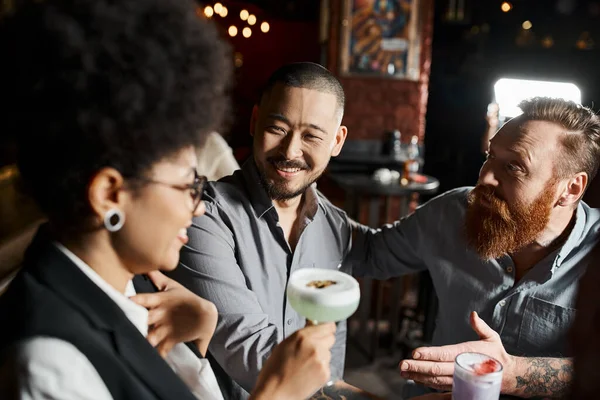 Бородатый татуированный мужчина разговаривает с удивленной африканской американкой рядом с азиатским коллегой в баре — стоковое фото
