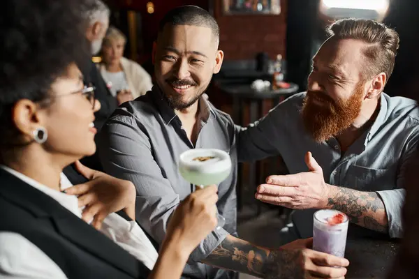 Barbuto tatuato uomo parlando con stupito africano americano donna vicino sorridente asiatico collega in bar — Foto stock