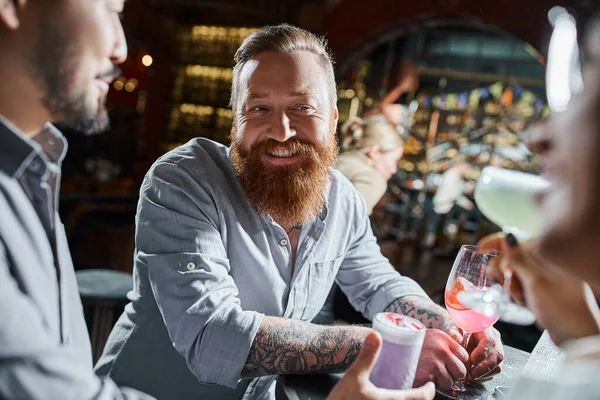 Homme barbu tatoué avec verre à cocktail souriant près de collègues multiethniques dans le bar, temps de fête — Photo de stock