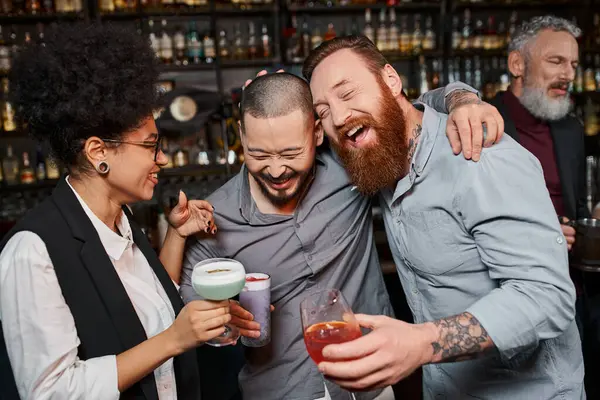 Colegas multiétnicos tatuados con cócteles riendo cerca de la alegre mujer afroamericana en el bar - foto de stock